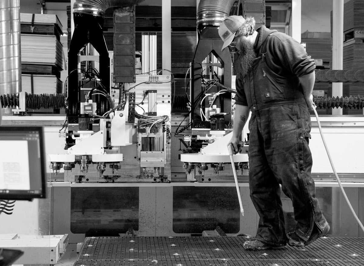 A black and white image of a man working at a CNC machine. He is blowing plastic shavings off of the machine deck.