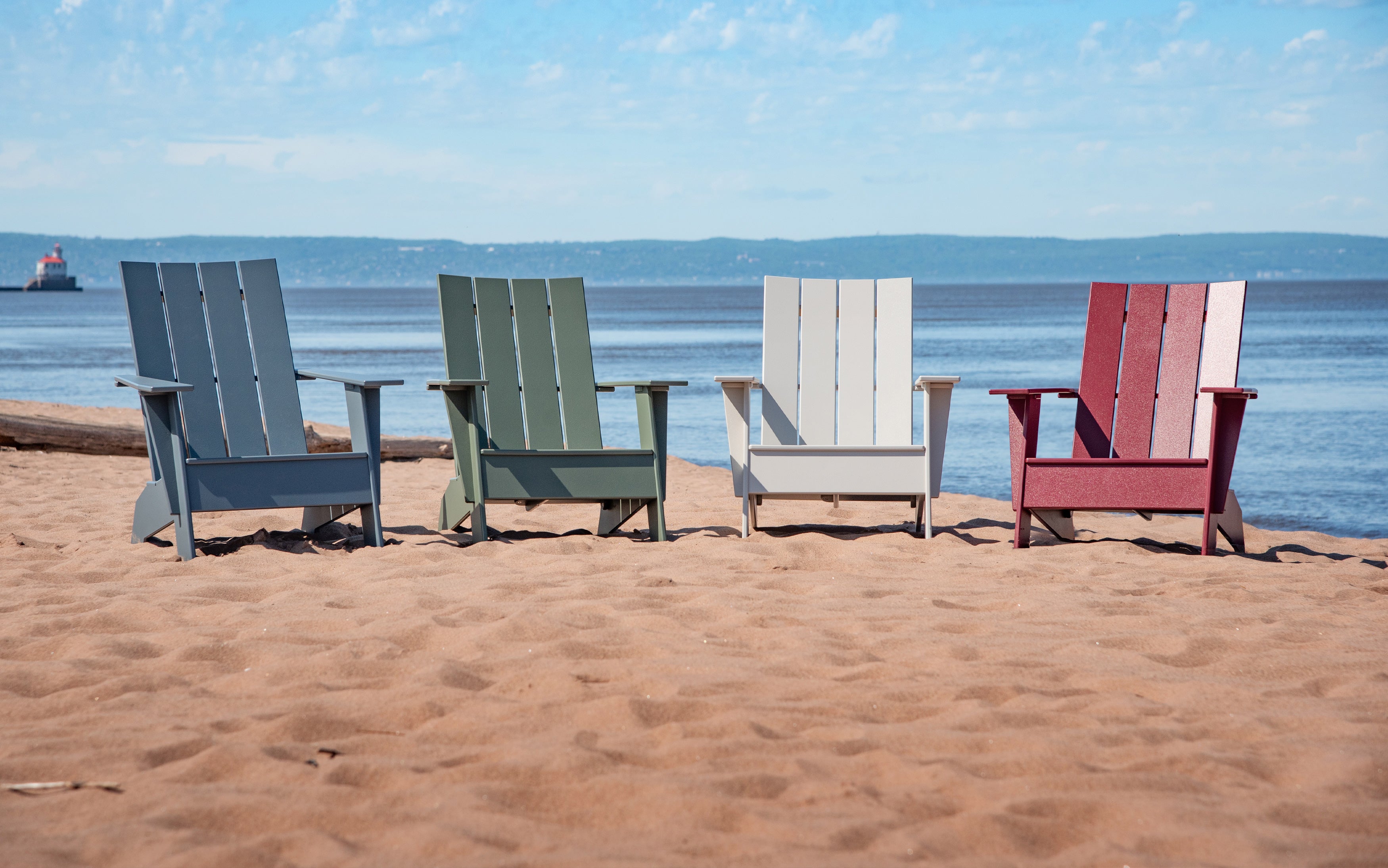 Four Adirondack Chairs in a row along a beach. From left to right, Ash Blue, Sage, Fog, and Chili.
