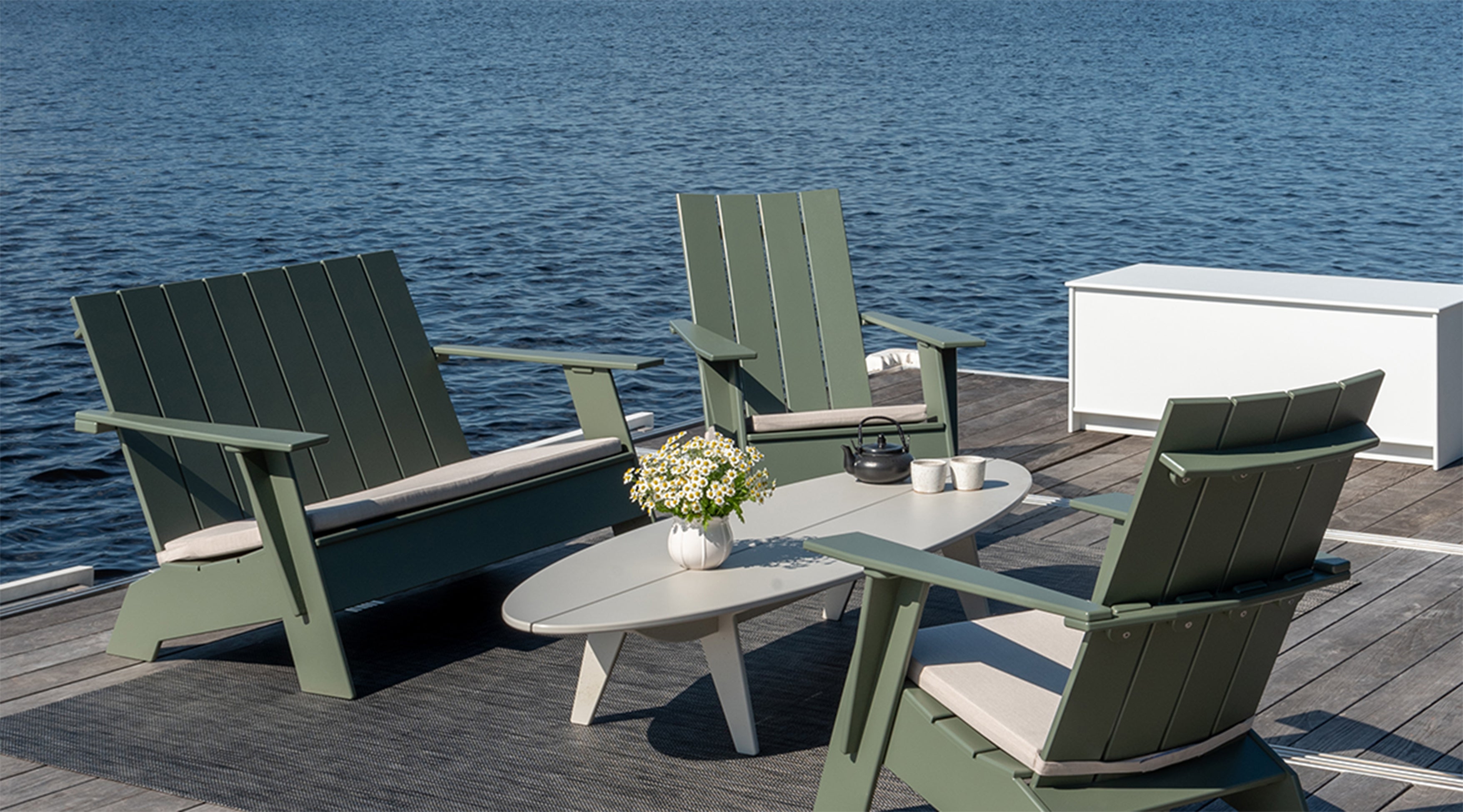 An Adirondack Bench and two Adirondack lounge chairs on a dock with a Bolinas Coffee Table with a Storage Chest and water in the background.