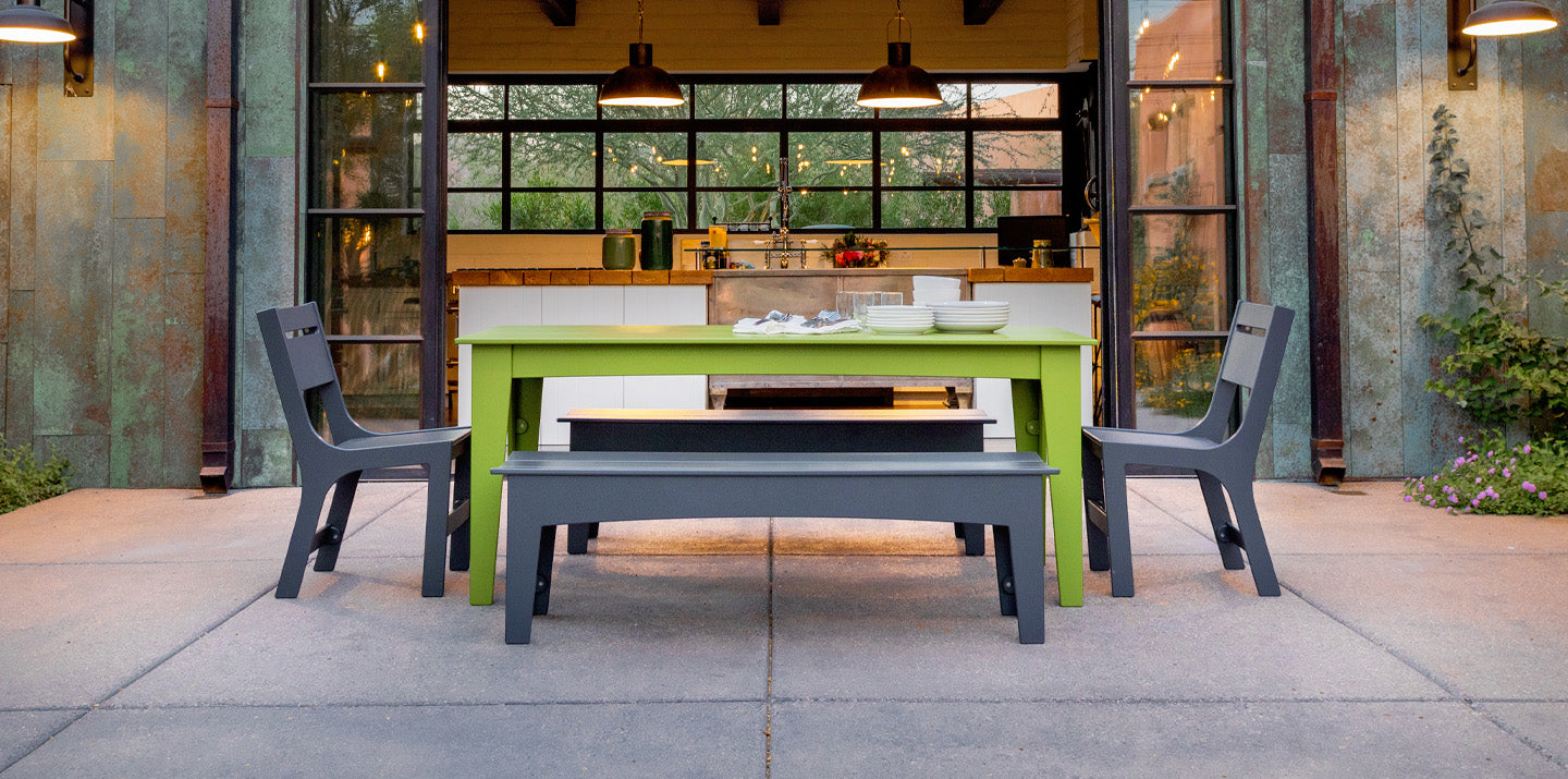 a stone patio with a leaf green alfresco table, charcoal alfresco dining chairs, and charcoal alfresco dining benchs. The back shows patio doors opened up to an indoor kitchen.