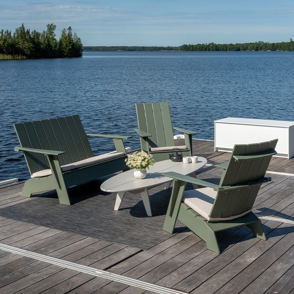An Adirondack Bench and two Adirondack lounge chairs on a dock with a Bolinas Coffee Table with a Storage Chest and water in the background.