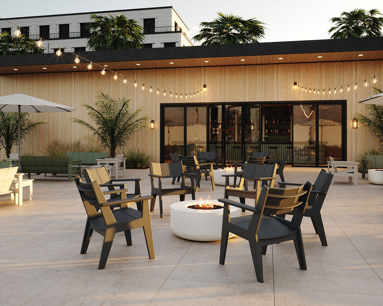 Outdoor patio seating area with black lounge chairs arranged around a round white fire pit, set in front of a wood-paneled building with large glass doors and string lights.