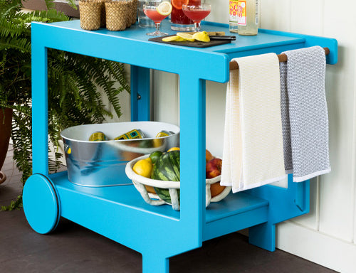 a sky blue lollygagger bar cart with various drink making materials staged on top. In the lower level of the bar cart, there is a small tub with cans and ice, along with a basket of fruit.