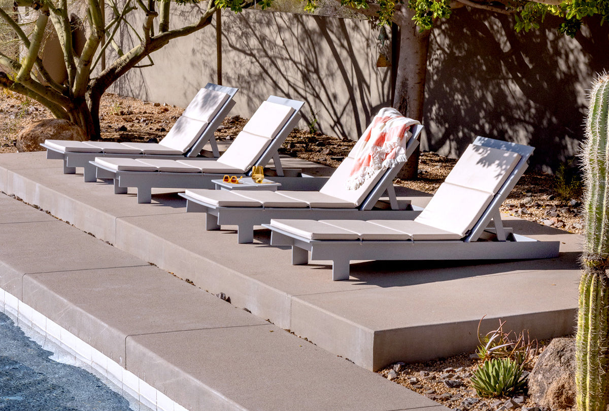 Four Driftwood Lollygagger Chaises along a pool with flax cushions on each product. A beach towel is draped over a chaise int he middle.