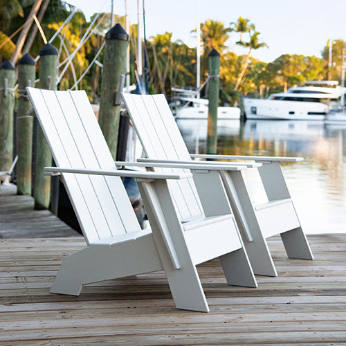 Two white Adirondack chairs on a wooden dock with boats and greenery in the background.