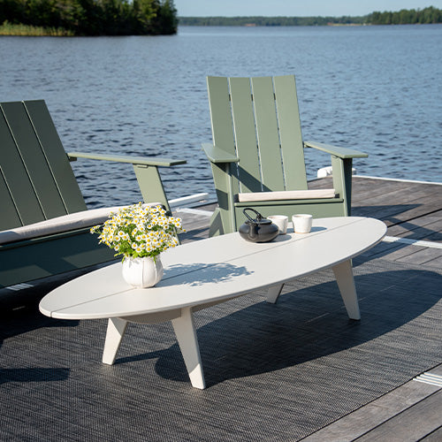 Two Adirondack chairs and a coffee table on a dock by a lake.