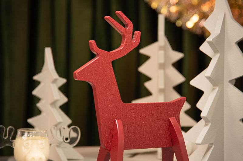 A red hdpe reindeer figurine stands on a tabletop in a styled holiday setting, surrounded by white geometric Christmas trees, a glass of eggnog, and warm blurred lights in the background.