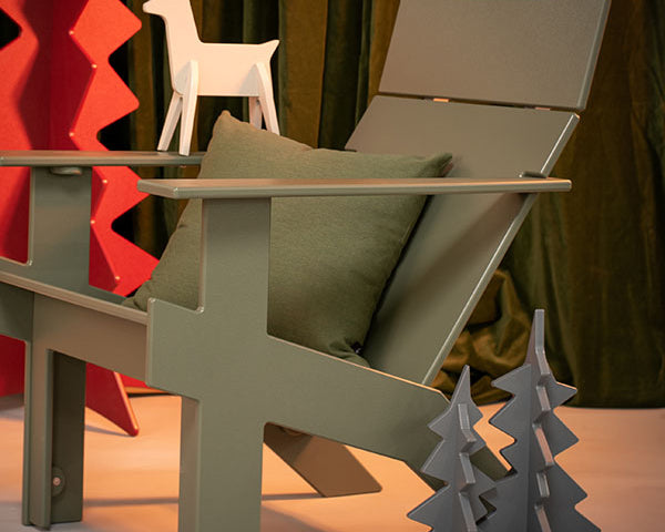 A modern sage green Adirondack-style chair with a matching green pillow, styled in a holiday-themed setting with a white reindeer figurine, red Christmas tree decor, and gray tree cutouts against a dark curtain background.