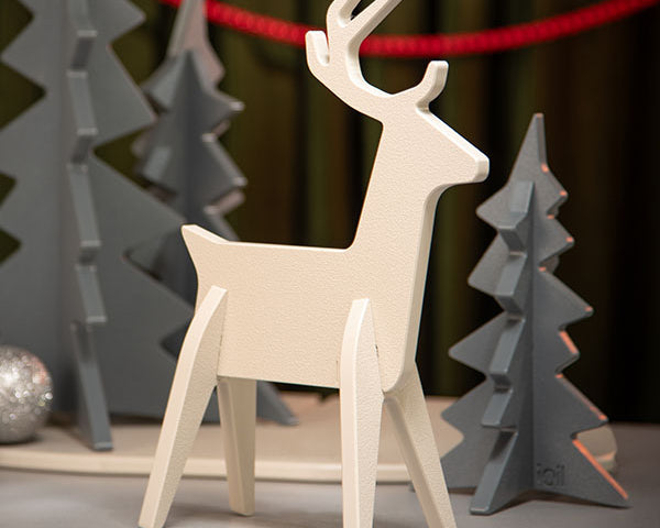 A minimalist white wooden reindeer figurine standing in front of gray geometric Christmas trees and a red garland, set against a dark green curtain background.