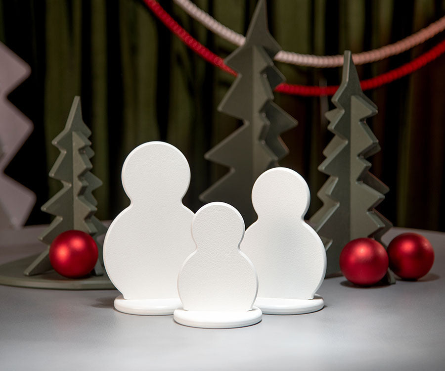 Three white snowman-shaped decorative figures of different sizes standing on a light gray surface, with gray tree silhouettes and red ornaments in the background.