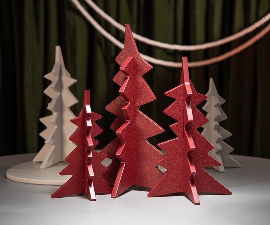 Three-dimensional geometric Christmas tree ornaments in red and off-white, arranged on a light gray surface with a dark green curtain backdrop.