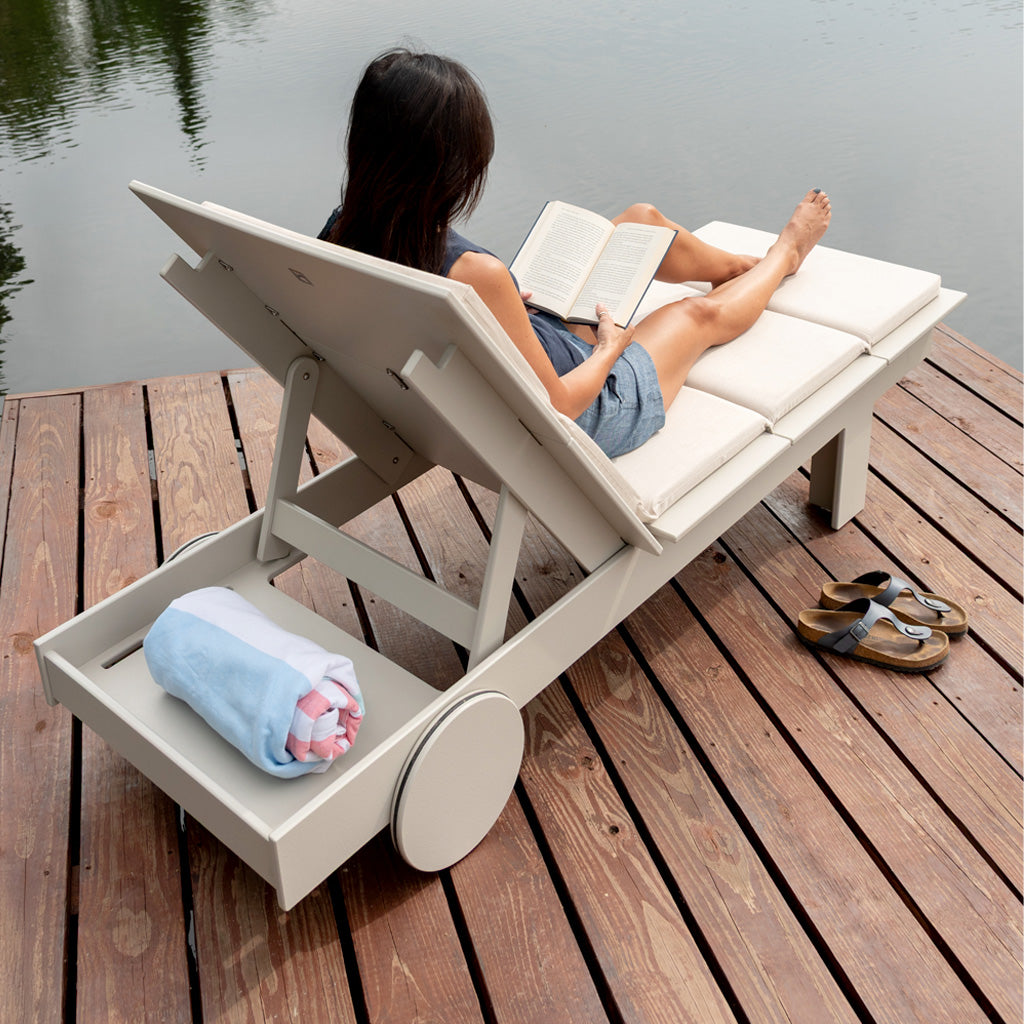 Image of a woman sitting in a tall chaise reading shot from behind chaise showing the shelf. A rolled towel is placed on the shelf. 
