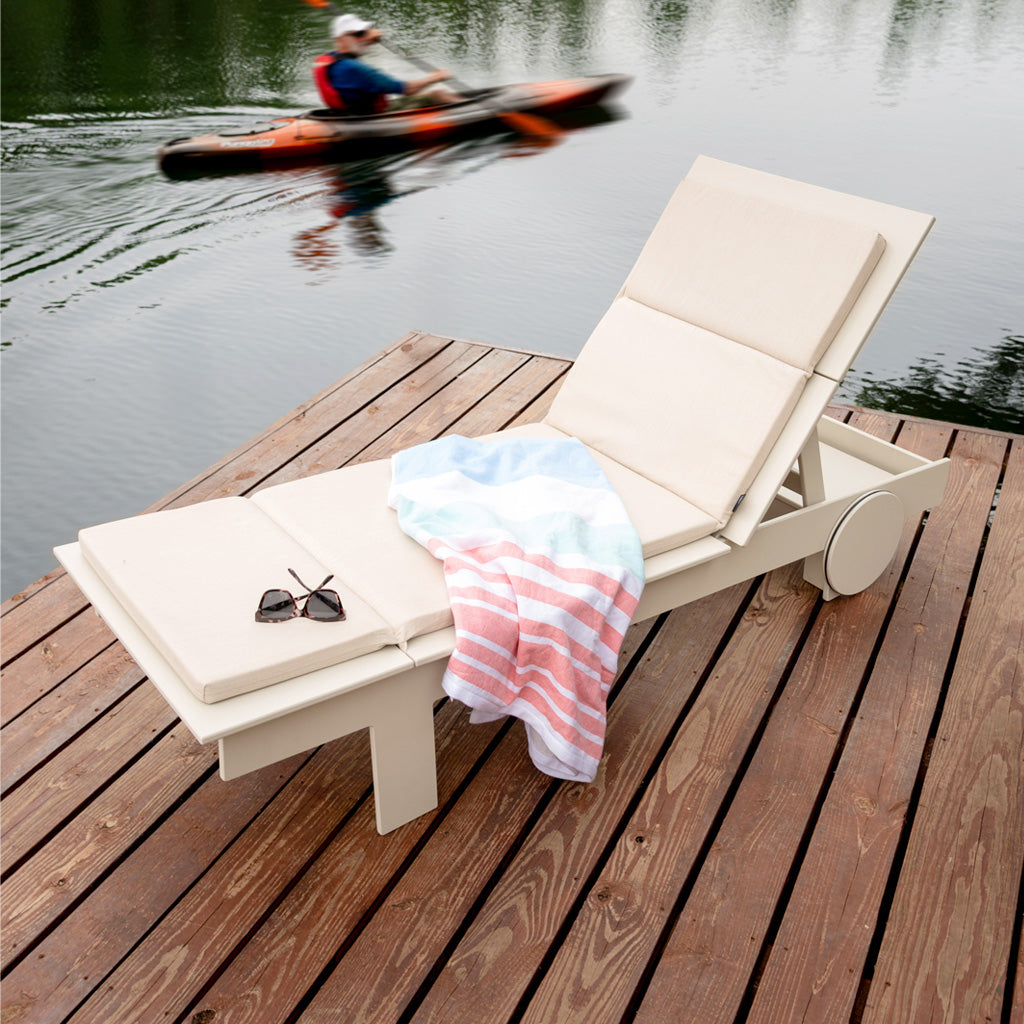 image of a fog tall chaise on a dock with a flax cushion. A towel and sunglasses are resting on the chaise. a man is paddling by in the lake in the background. 